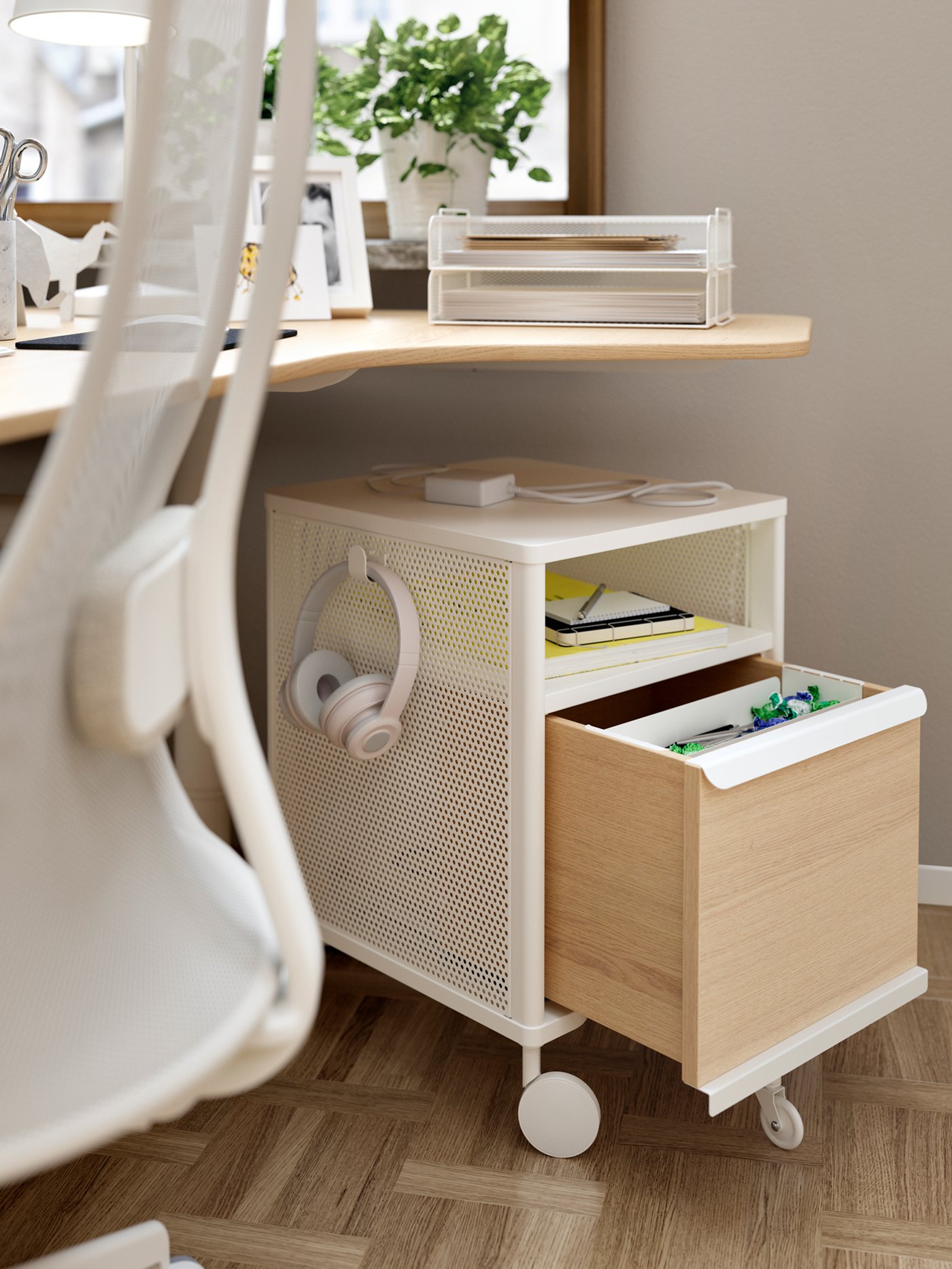 A white-mesh BEKANT storage unit on castors standing on the parqueted floor beneath a BEKANT sit-stand corner desk.