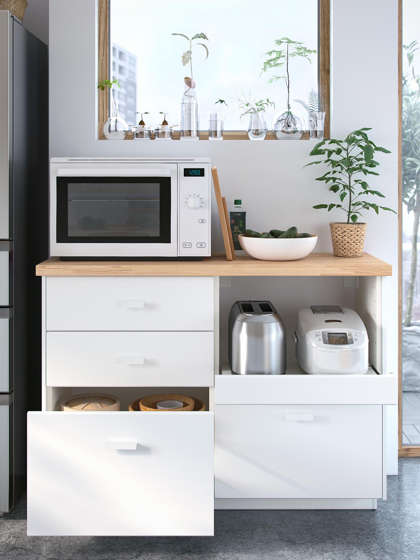 A white microwave stands below a window on top of a wooden worktop with four white drawers below, one is open.