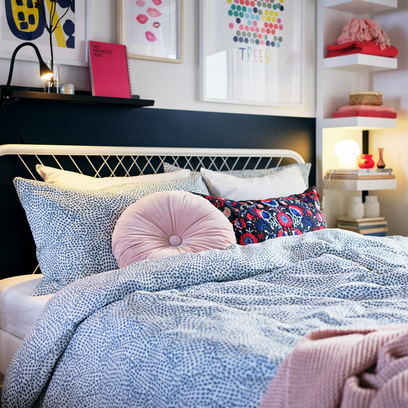 A white NESTTUN bedframe covered in a duvet with a pink throw and round cushion with art and open shelves on the wall behind.