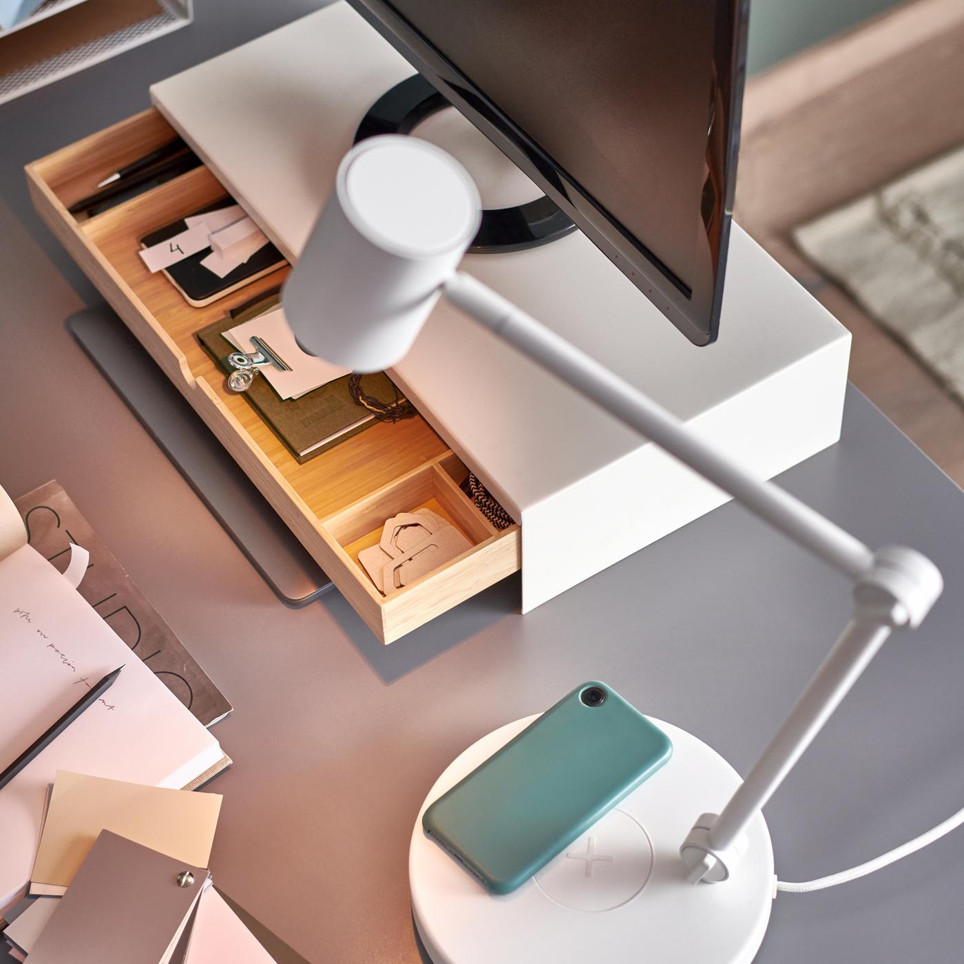 A white NYMÅNE LED work lamp with wireless charging, plus a computer screen on a monitor stand with an open drawer.