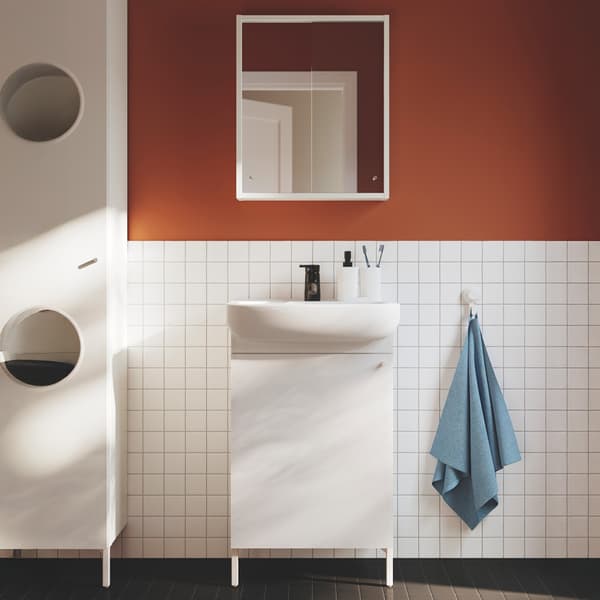 A white NYSJÖN wash-stand with door with a white BJÖRKÅN wash-basin with tap leaned against the  white square tile and a white framed mirror mounted on the orange/red wall. A white NYSJÖN
laundry cabinet is seen on the left side of the bathroom, while a light blue towel is hung on the right of the wash-stand.