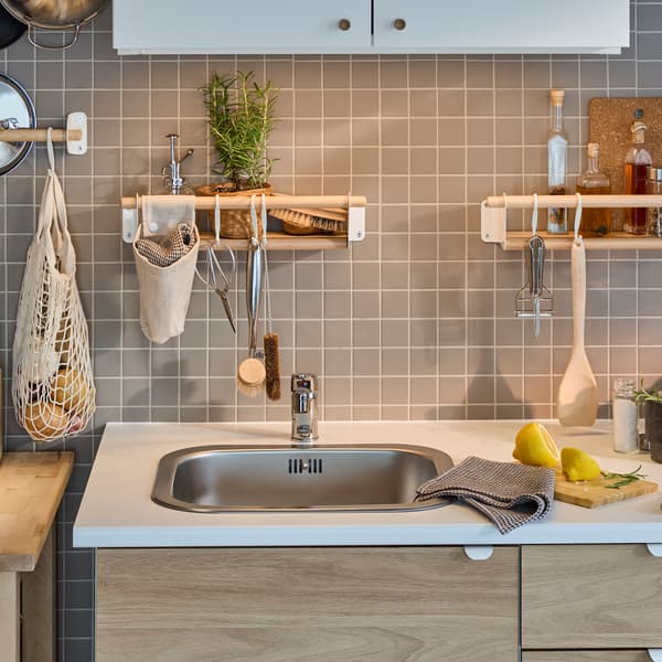 A white/oak effect ENHET kitchen by a grey-tiled wall with a birch rail, two birch wall shelves and a white wall cabinet.