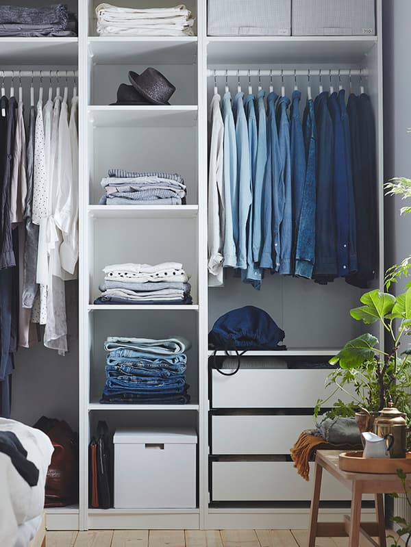 A white PAX wardrobe containing a lot of clothes including hats and folded clothes on shelves and shirts hanging from rails.