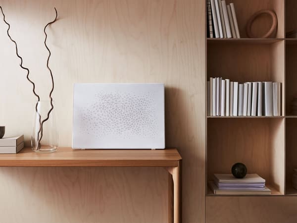 A white picture frame with a built in speaker sitting on a wooden desk with a wooden wall behind and a bookshelf next to it.