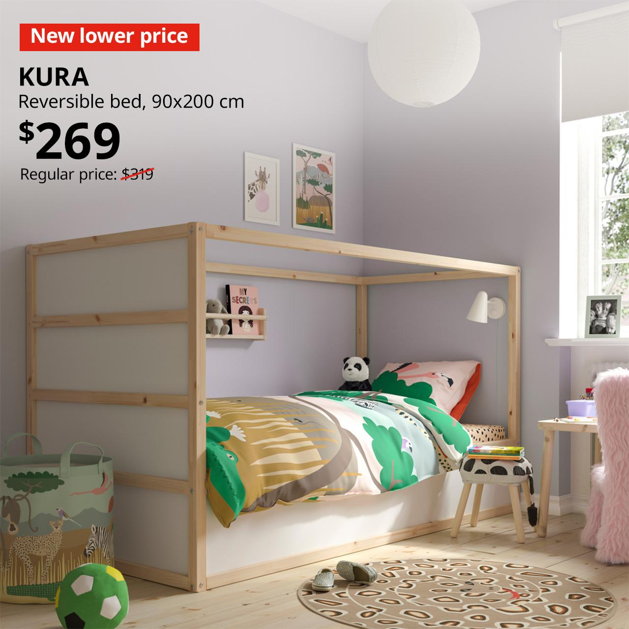 A white/pine KURA reversible bed in a children's room setting, featuring a light purple wall with playful decorations. A red tag with white text that reads "New lower price", followed by a black text overlay image that reads, "KURA, Reversible bed, 90x200 cm, 90x200 cm, $269, Regular Price: $319".