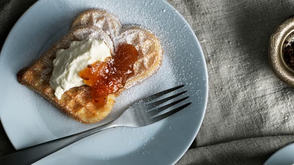 A white plate with a fork and two waffles with whipped sour cream and cloudberry jam. Powdered sugar is sprinkled on top.