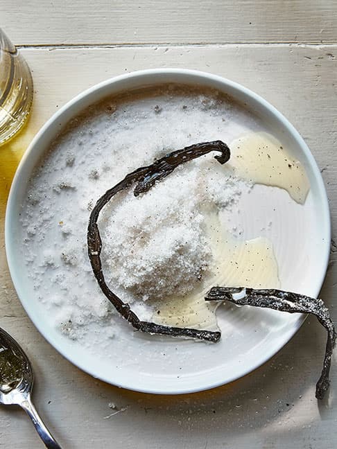 a white plate with a heap of white sugar and two vanilla pods on it, next to a spoon with some liquid and a glass jar with a yellowish liquid in the background