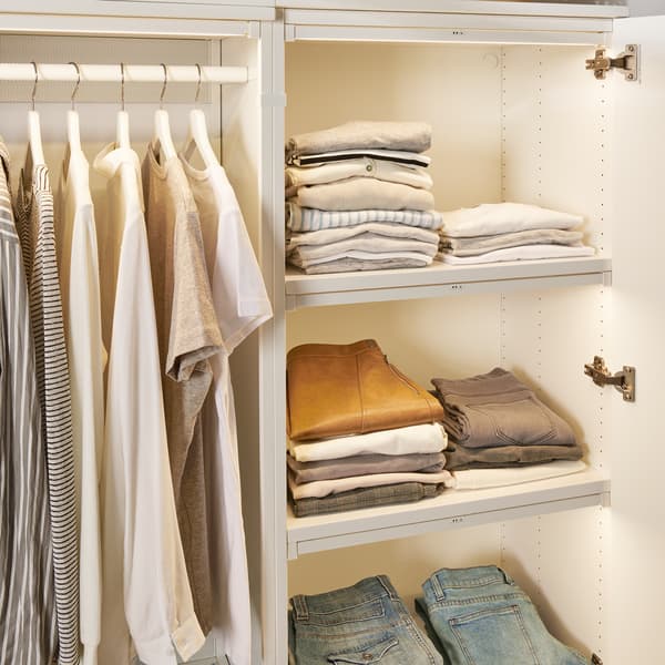 A white PLATSA cabinet with lighting. There are piles of clothes in the closet and shirts hanging on the rod. 