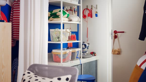 A white PLATSA open wardrobe with dark grey LARKOLLEN sliding doors near a door, holding storage boxes and folded clothes.