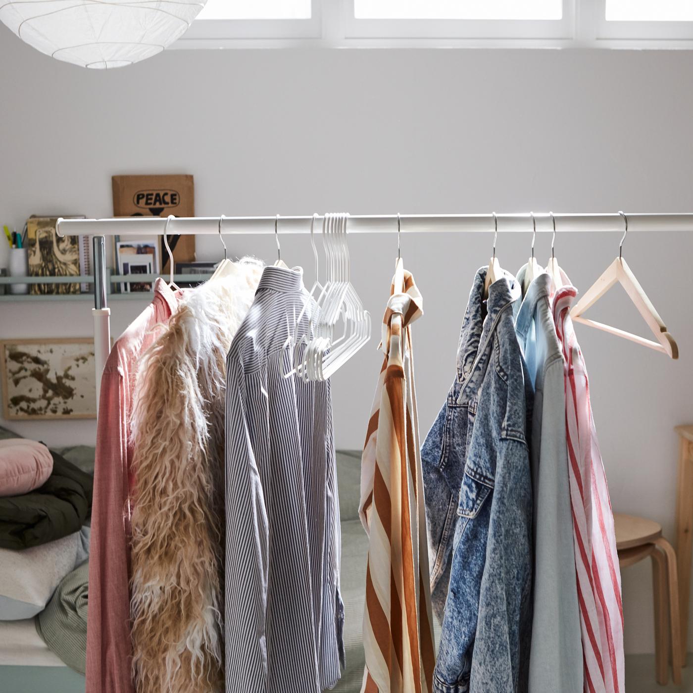 A white RIGGA clothes rack with clothes on hangers beside a pine table and a bed with various cushions and bedding in a pile.