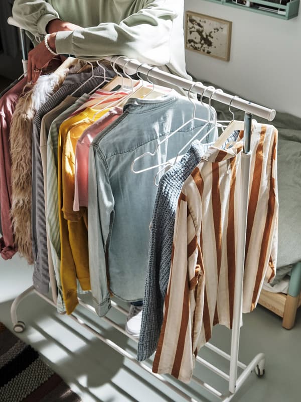 A white RIGGA clothes rack with several shirts in different colours and a young person in a green sweater leaning on it.
