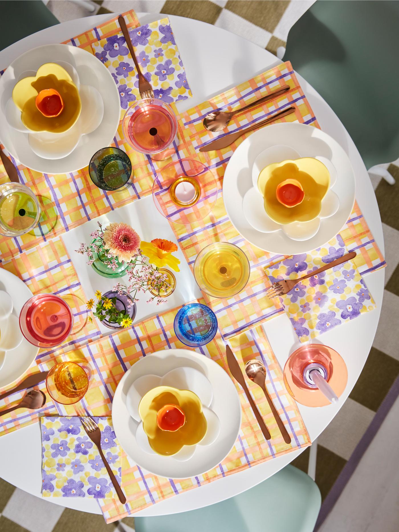 A white, round table seen from above is laid for four persons with placemats, tableware and glassware in vibrant colours.