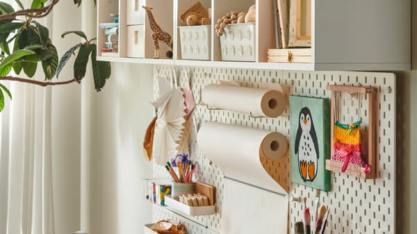 A white shelf, containing children’s toys and boxes, hangs above a SKÅDIS pegboard with paper rolls and pens.