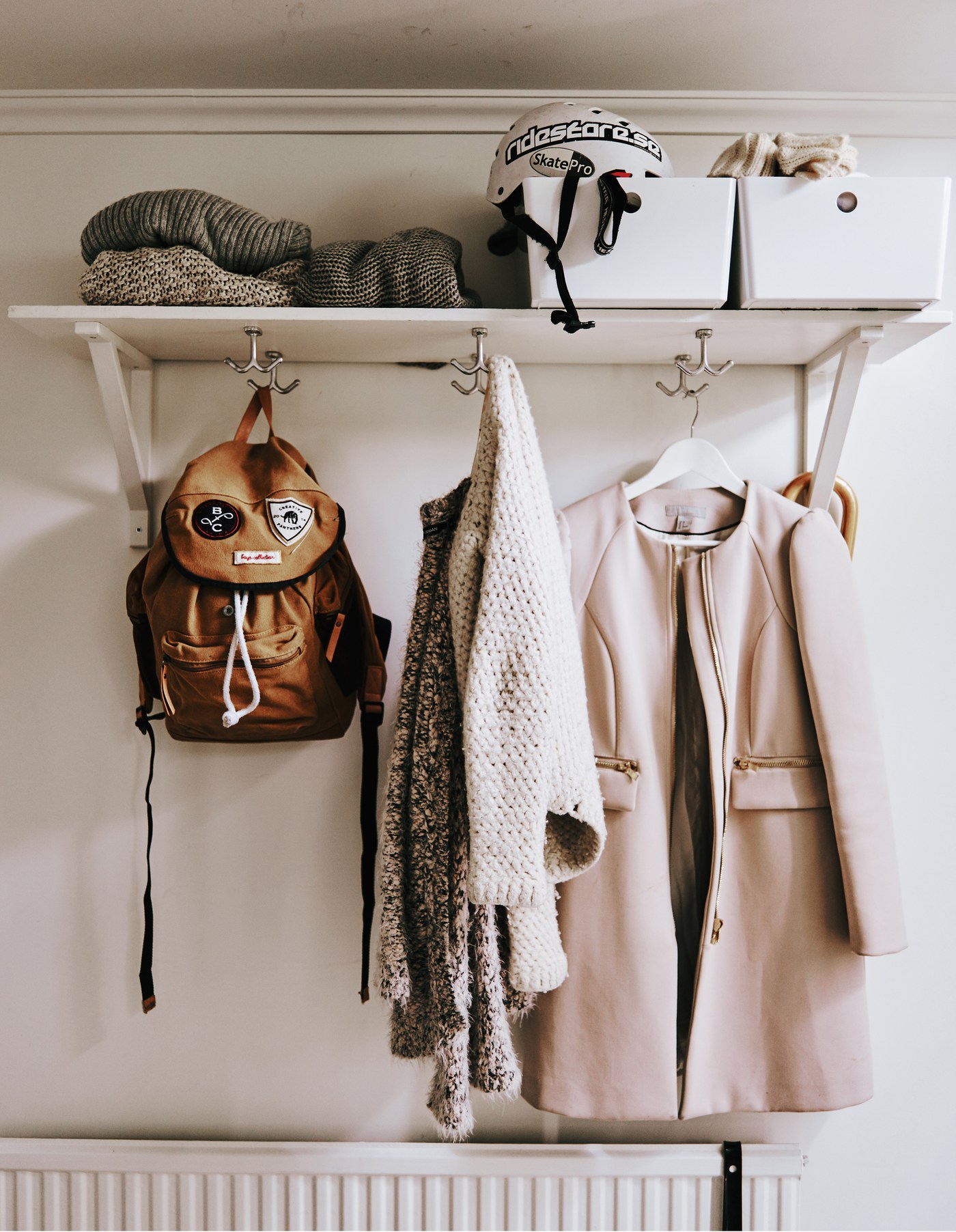 A white shelf mounted high up on the wall in a hallway with hooks on the bottom shelf to hang coats, backpacks and coats.