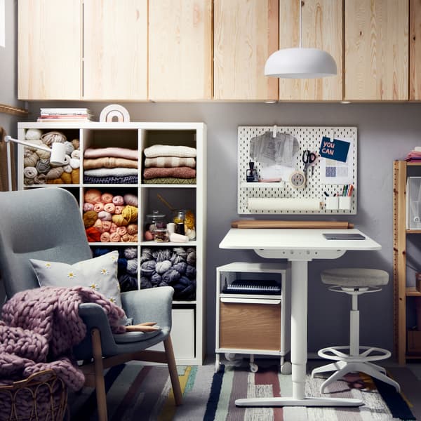A white shelving unit with yarn, a light green armchair, a white desk with sit/stand support, pine cabinets and a pendant.