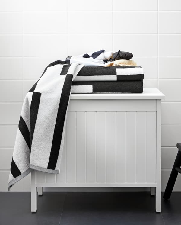 A white SILVERÅN storage bench with towels with a graphic black and white pattern on top of it, standing in a bathroom.