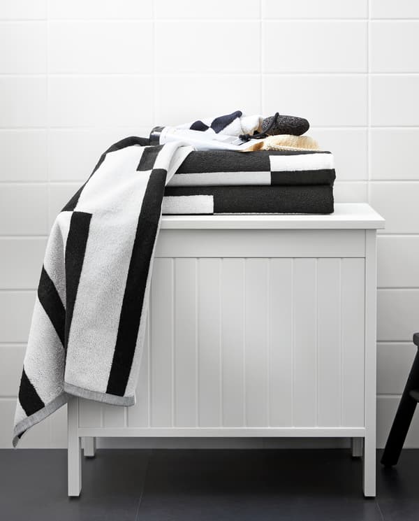 A white SILVERÅN storage bench with towels with a graphic black and white pattern on top of it, standing in a bathroom.
