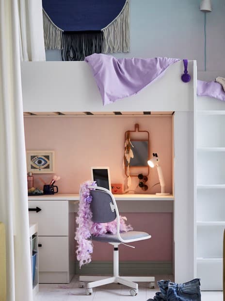 A white SMÅSTAD loft bed with a desk that has an ÖRFJÄLL swivel chair in front of it with a feather boa draped over the back.