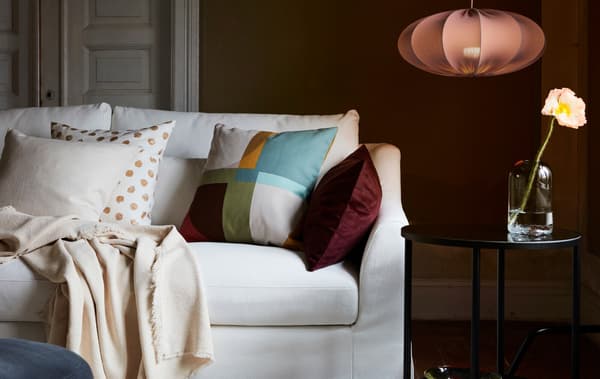 A white sofa in a living room with a mix of cushions and a throw. Next to it is a black side table and REGNSKUR lamp.