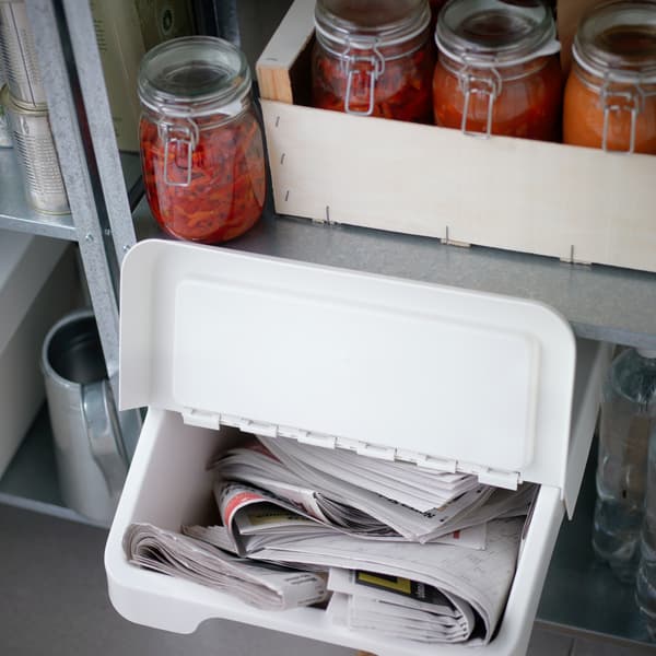 A white SORTERA waste sorting bin with an open lid containing newspapers, placed under a shelf with jars of pickled red vegetables