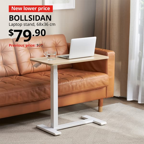 A white stained oak BOLLSIDAN laptop stand in a the living room setting right next to a LANDSKRONA 3-seat sofa. A red tag with white text that reads "New lower price", followed by black text over image that reads, "BOLLSIDAN, Laptop stand, 68x36 cm, Price: $79.90. Previous price with red slash: $99".