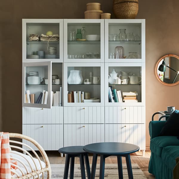A white storage combination with glass-doors and drawers, a round mirror in rattan and a dark blue-green nest of two tables.