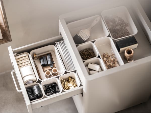A white storage unit with an open drawer and open top filled with different sized beige NOJIG boxes holding small objects.