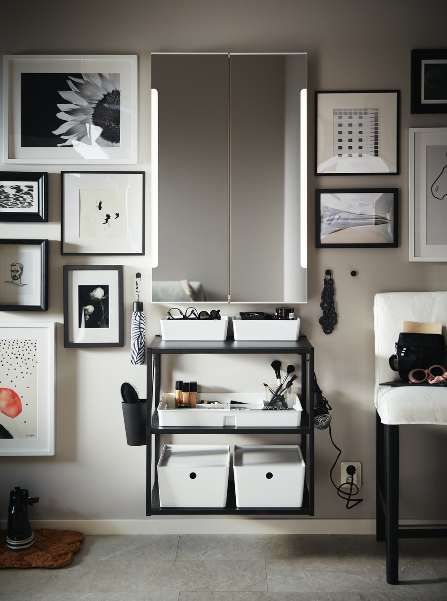 A white STORJORM mirror cabinet with built-in lighting on a wall with framed pictures and an anthracite shelving unit.