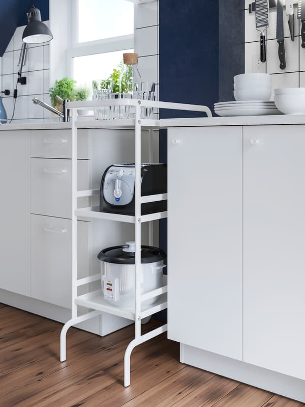 A white SUNNERSTA kitchen trolley holding a rice cooker and toaster stands in a gap between two white kitchen cabinets.