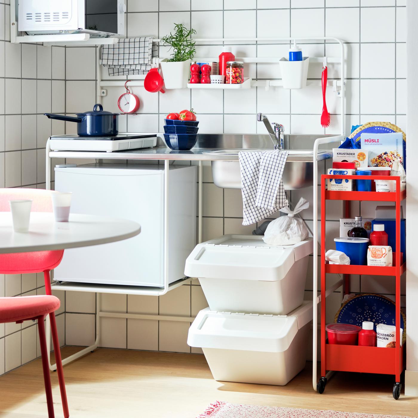 A white SUNNERSTA mini-kitchen, kitchen appliances, waste sorting bins, a red-orange trolley and a table with a red chair.