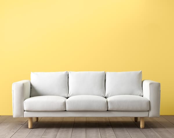 A white three seat sofa on a wooden floow with a yellow background behind it. 