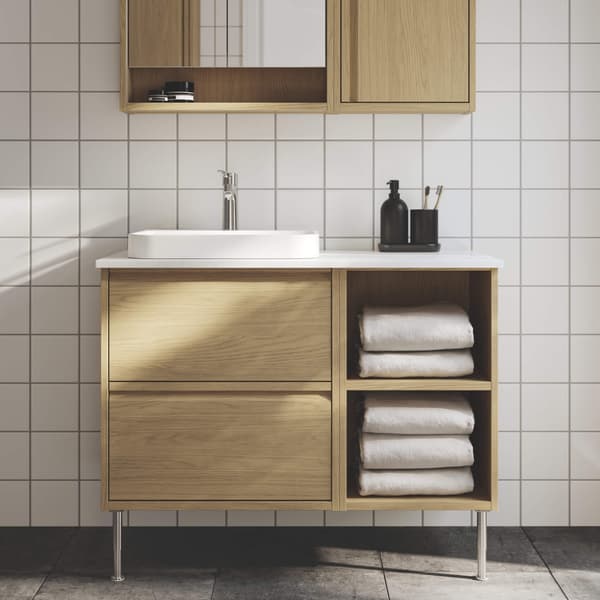 A white-tiled bathroom has an ÄNGSJÖN sink cabinet in oak effect with legs, a semi-recessed sink cabinet and a mirror cabinet.