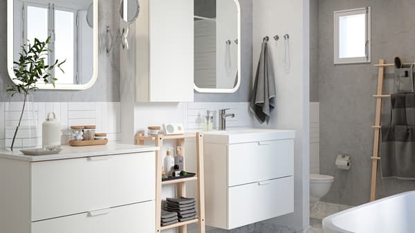 A white-tiled bathroom with grey walls, two wash-stands, mirrors and a shelf in birch with towels and beauty products.