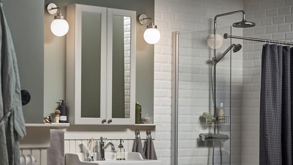 A white-tiled bathroom with light green walls, two FRIHULT wall lamps on either side of a GODMORGON mirror cabinet.