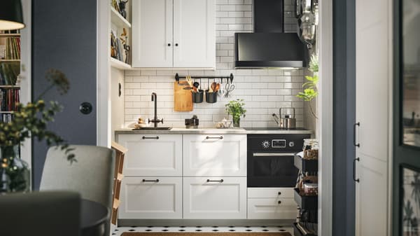 A white-tiled kitchen with matte white AXSTAD fronts and tall cabinets has black appliances and shelves on the wall.