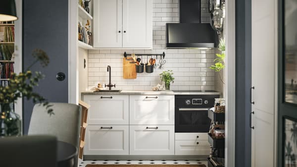 A white-tiled kitchen with matte white AXSTAD fronts and tall cabinets has black appliances and shelves on the wall.