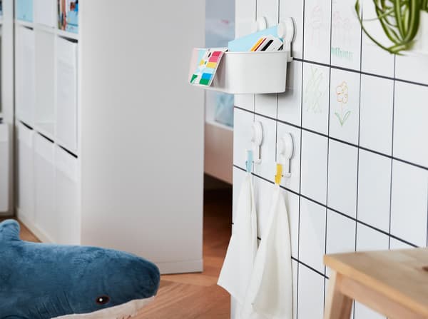 A white tiled wall with TISKEN basket and hooks with suction cups attached to it, they hold children's toys and small towels.