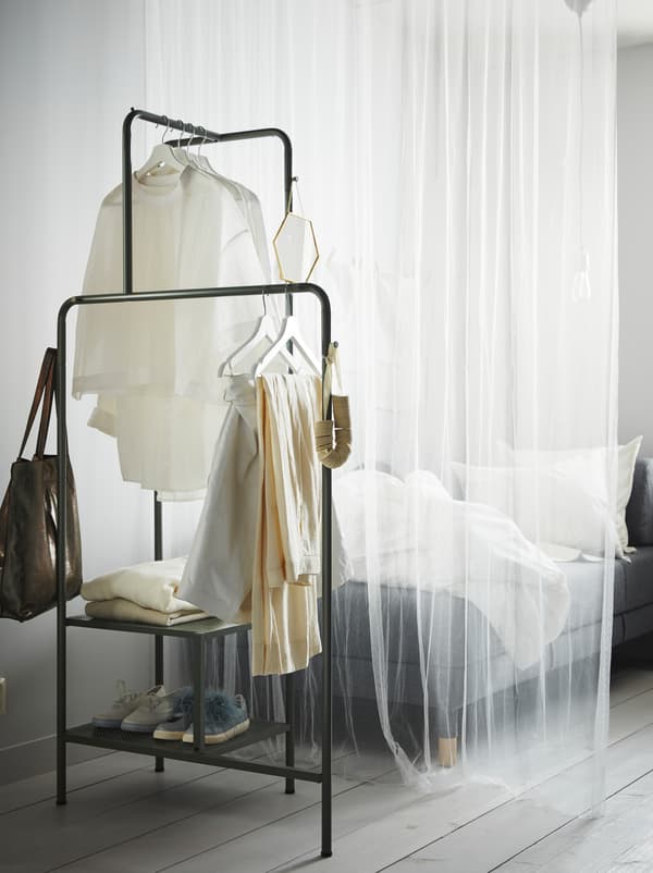 A white, transparent curtain separates a sofa from a grey-green NIKKEBY clothes rack with shoes, clothes, and accessories.