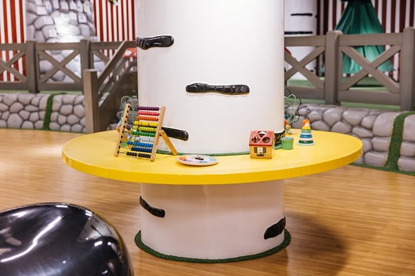A white tree-like column with a circular yellow play table wrapped around it, featuring colorful educational toys such as an abacus, stacking rings, and a toy house, set on a wooden floor in a forest-themed children's play area.