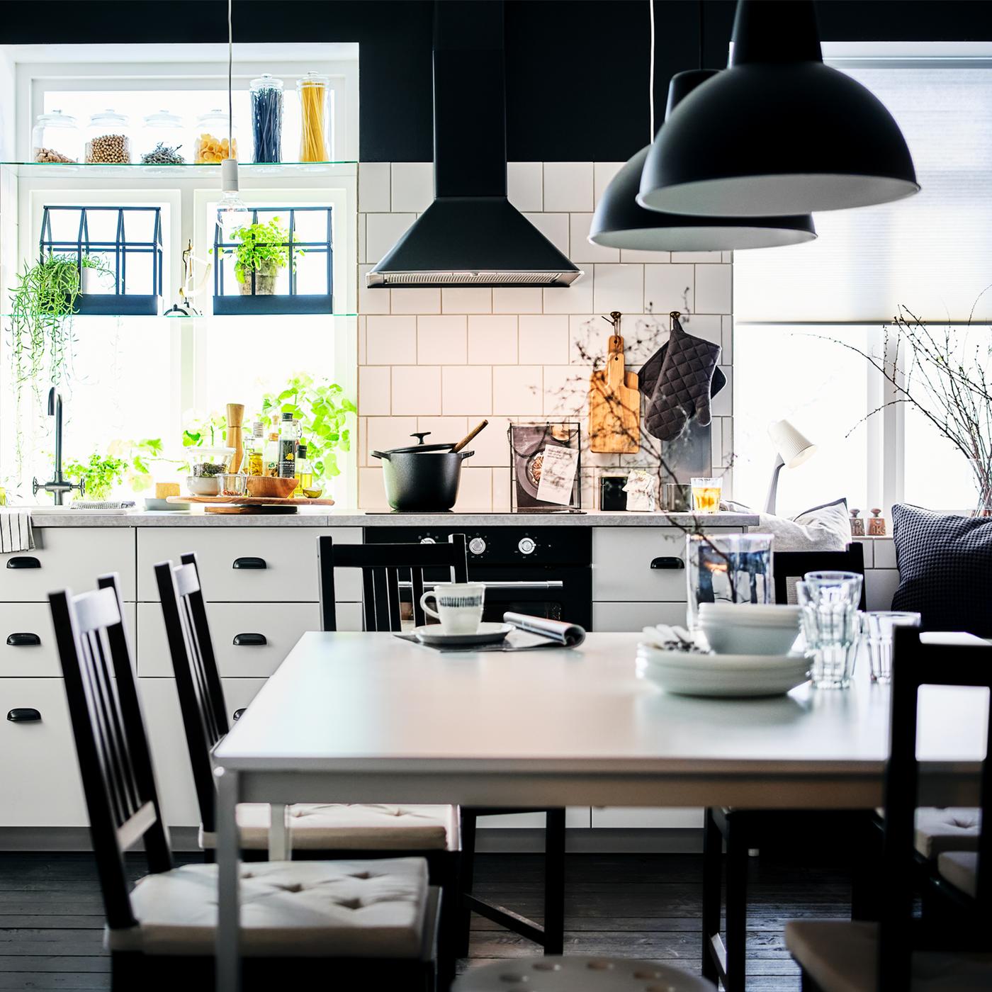 A white VEDDINGE kitchen with five brown-black STEFAN chairs at white MELLTORP tables, under black SKURUP pendant lamps.