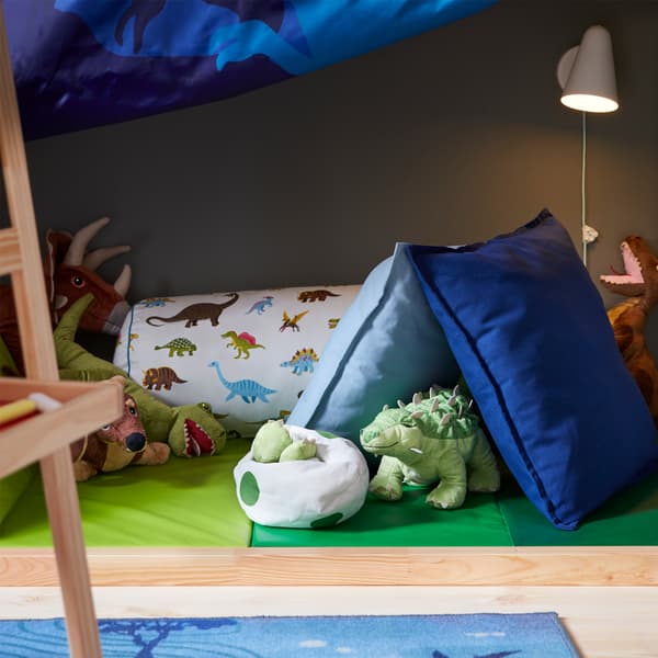 A white wall lamp that’s lit and a green folding gym mat with dinosaur soft toys and blue/dinosaur-patterned cushions.