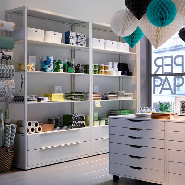 A white-walled paper shop with colourful paper lanterns, white shelving units and white ALEX drawer units on castors.