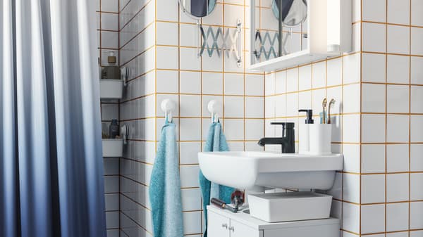A white wash-basin cabinet, a black tap, white tiles with yellow grout, a dark blue shower curtain, hooks with towels.
