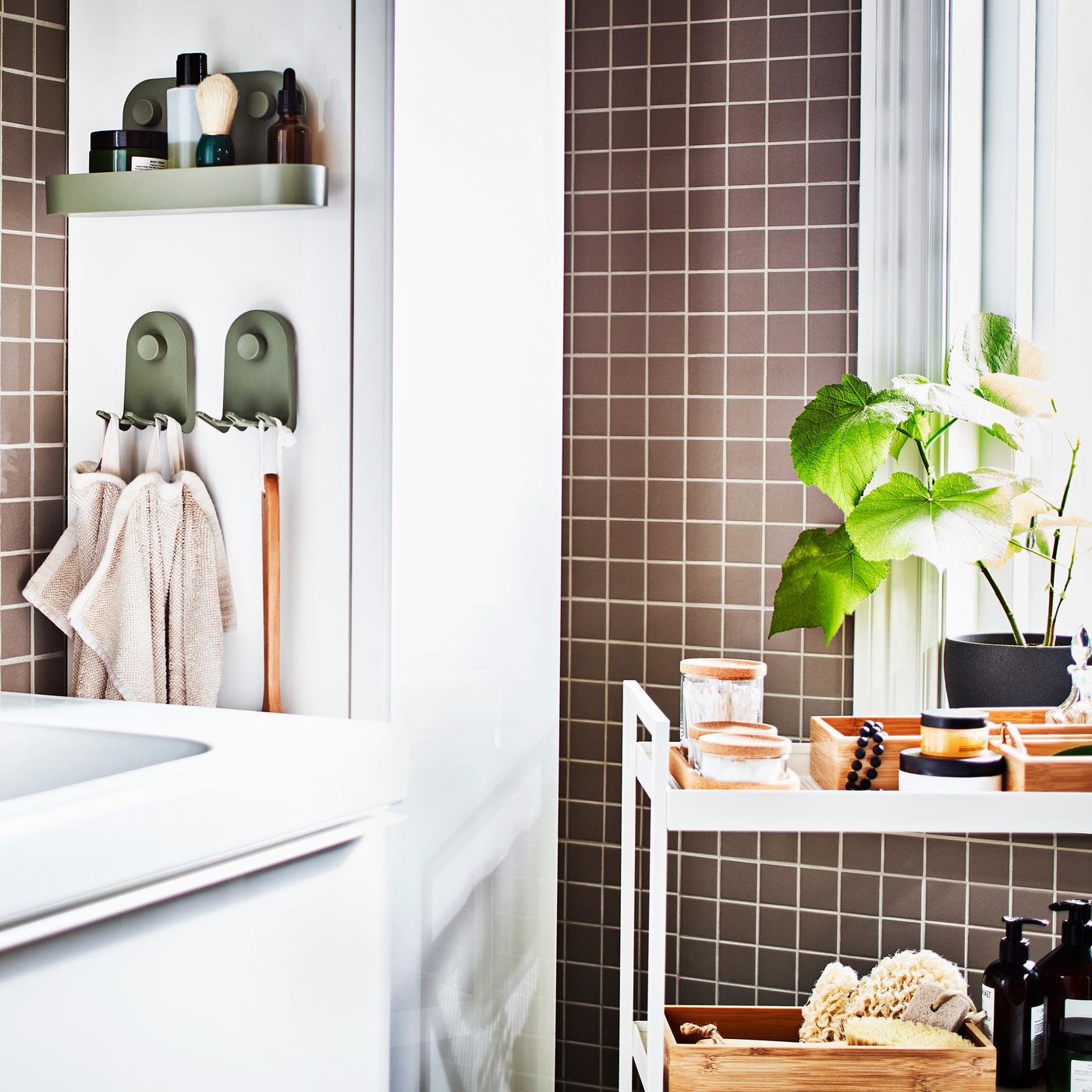 A white wash-stand, a white trolley with trays and jars, a white high cabinet and a potted plant in a grey-tiled bathroom.