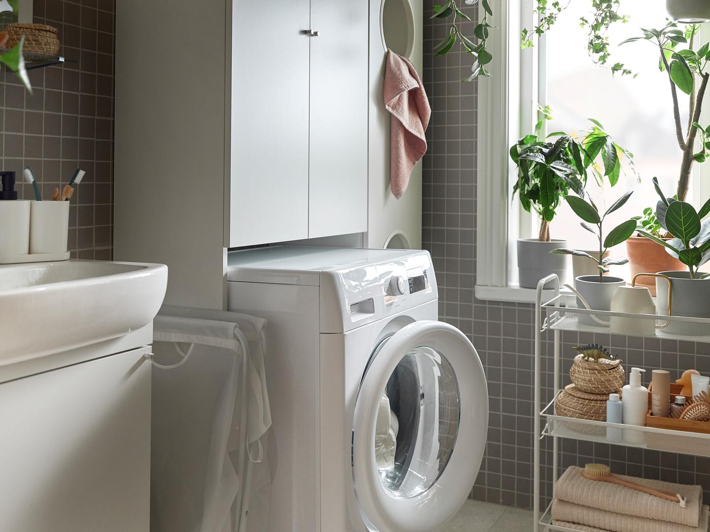 A white washing machine in a bathroom, placed inside ENHET laundry solution. Plants in the window and a trolley next to it.