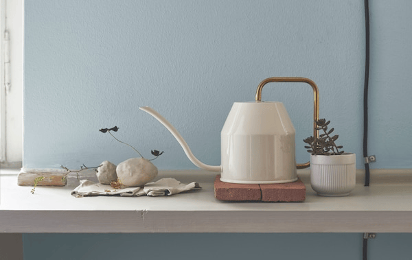 A white watering can with gold-colour handle on a shelf with pot plant and stones.