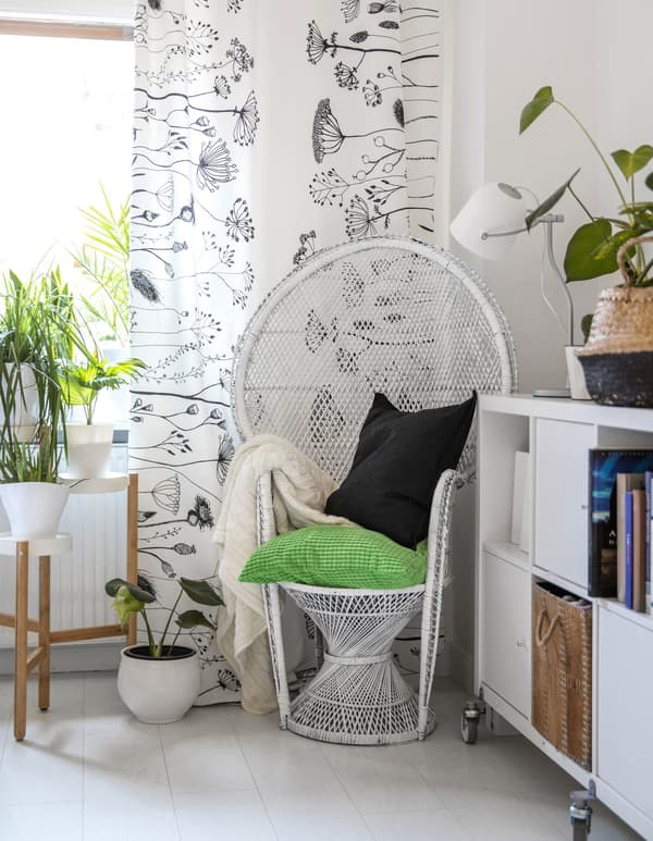 A white wicker chair in the corner of a white room with green touches and plants.
