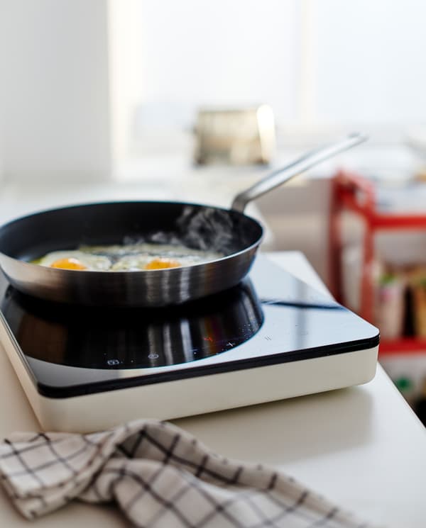 A white worktop with a black/white portable induction hob, a black/white tea towel and a frying pan where eggs are fried.