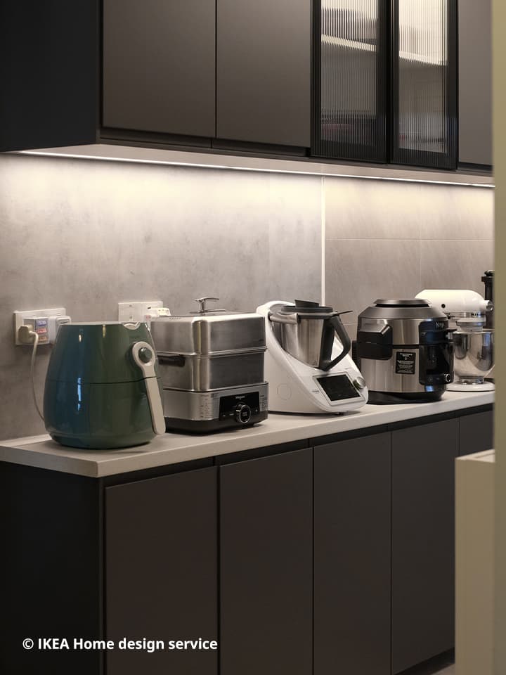 A wide view of an IKEA METOD kitchen system with grey door fronts and cabinets, installed with integrated lighting. On the kitchen counter sits various appliances such as an air fryer, mixer, steamer, pressure cooker and other kitchen equipment.