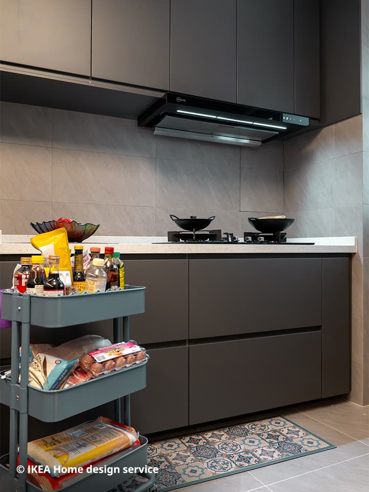 A wide view of an IKEA METOD kitchen system with grey door fronts and cabinets, along with the hob and hood extractor. On the bottom left corner sits a grey-blue RÅSKOG trolley filled with various sauces and seasonings.